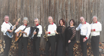 Eine Gruppe von Musikern, gekleidet in passenden Outfits, posiert für ein Foto vor einer Holzscheune mit einem Stapel Brennholz dahinter.