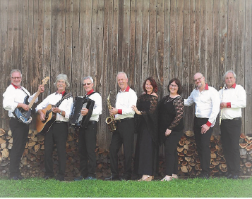 Eine Gruppe von Musikern, gekleidet in passenden Outfits, posiert für ein Foto vor einer Holzscheune mit einem Stapel Brennholz dahinter.