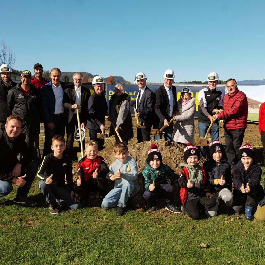 Eine Gruppe von Menschen in Hüten und Jacken, einige mit Schaufeln, posiert für ein Foto an einer Baustelle. Erwachsene und Kinder sind im Vordergrund und Hintergrund.