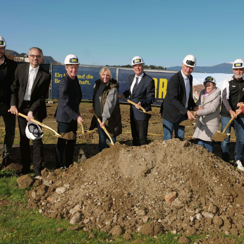 Eine Gruppe von Personen mit Schutzhelmen und Schaufeln steht um einen Haufen Erde herum, vermutlich an einer Baustelle.