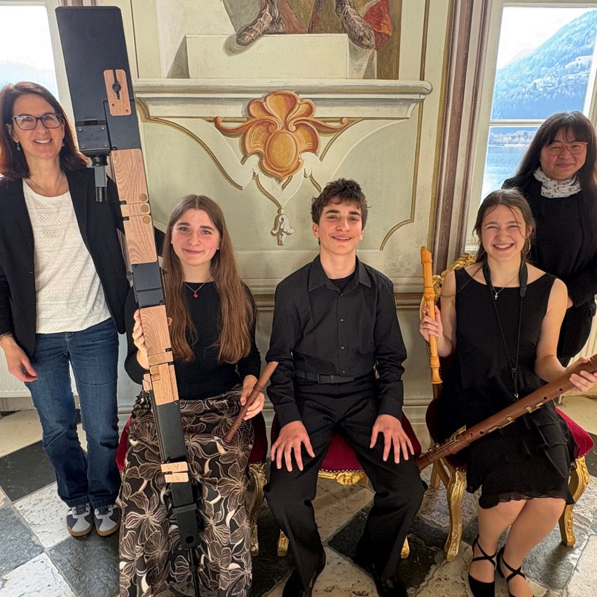 Fünf Personen posieren drinnen für ein Foto, drei halten lange Holzinstrumente. Die Umgebung ist mit kunstvollen Dekorationen geschmückt und bietet einen Blick durch ein Fenster.