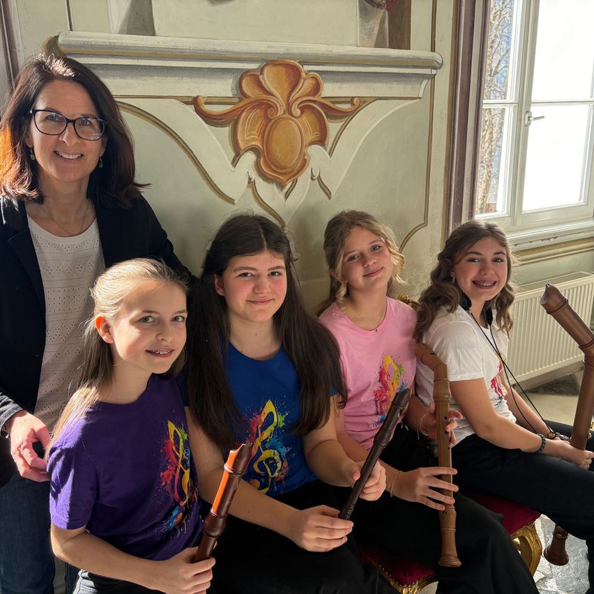 Eine Frau mit Brille und Halskette steht hinter vier Mädchen, die Holzflöten halten. Die Mädchen lächeln und sitzen auf einem Stuhl. Dahinter befindet sich eine dekorative Wand mit einem Blumendesign.