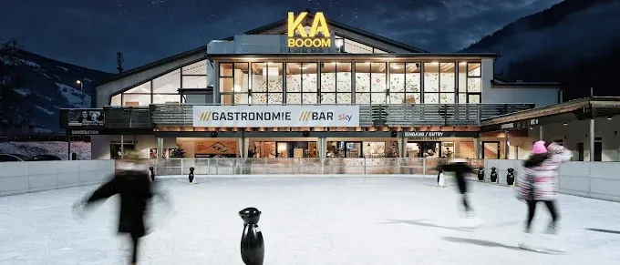Eine Eisbahn mit mehreren Menschen beim Schlittschuhlaufen. Ein Gebäude mit dem Schild KA BOOM darüber. Das Gebäude hat einen Bar- und Gastronomiebereich.