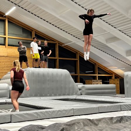 Eine Frau springt über ein aufblasbares Hindernis, während vier Männer zusehen. Die Szene spielt sich drinnen ab, mit Holzwänden und einer weißen Decke.