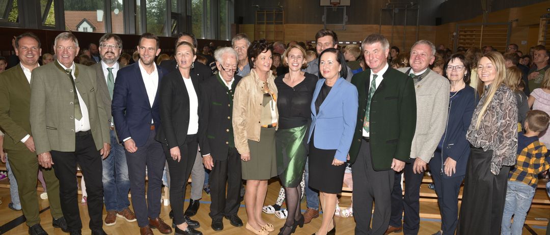 Eine Gruppe von Menschen steht zusammen in einer Turnhalle, lächelt und posiert für ein Foto. Dahinter ist ein Basketballkorb zu sehen, und der Raum ist mit großen Fenstern gut beleuchtet.