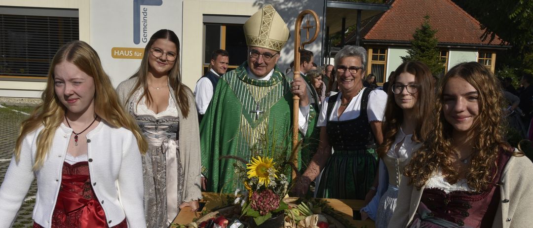 Ein Priester steht an einem Tisch mit einer Obstanordnung, umgeben von lächelnden Frauen in traditioneller Kleidung. Ein Gebäude mit einem Schild, das 'HAUS fur Gemeinde' liest, ist im Hintergrund sichtbar.