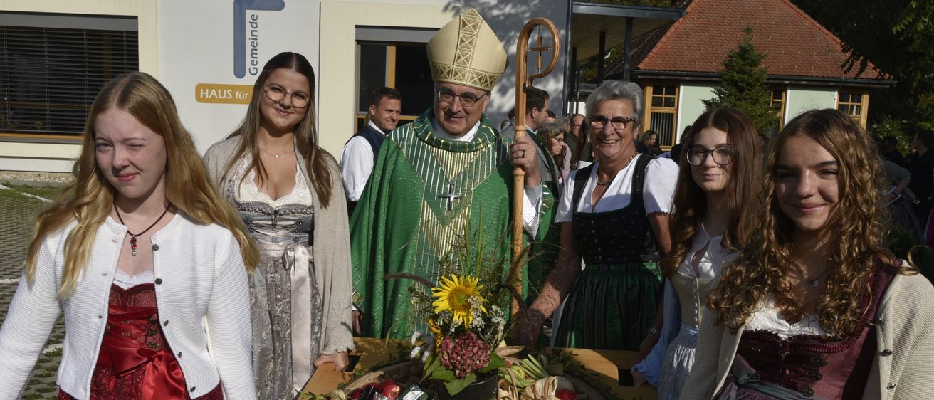 Ein Priester steht an einem Tisch mit einer Obstanordnung, umgeben von lächelnden Frauen in traditioneller Kleidung. Ein Gebäude mit einem Schild, das 'HAUS fur Gemeinde' liest, ist im Hintergrund sichtbar.