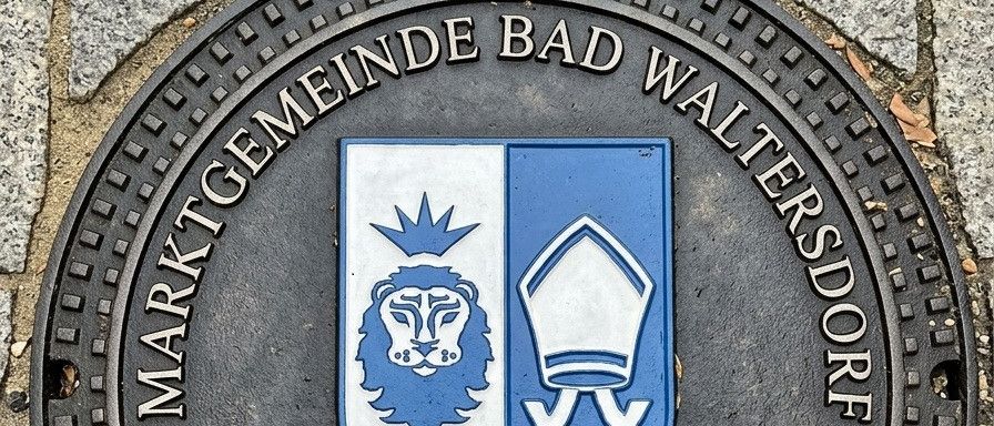 Eine schwarze runde Plakette mit einem Löwen, einer Krone und Text auf Deutsch. Es steht 'Marktgemeinde Bad Waltersdorf'. Darunter 'Digitalisierung Kanalsystem ab April 2026'.