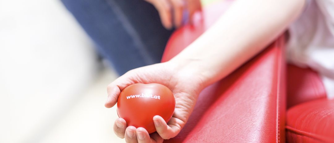Eine Hand hält einen roten Ball mit der Aufschrift www.blut.at, während eine andere Person in Blau sich vorbereitet, Blut abzunehmen.