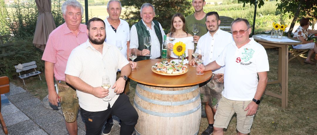 Acht Personen posieren für ein Foto in einer Außenumgebung. Sie halten Weingläser, mit einer Pizza auf einem Fass in der Mitte. Der Hintergrund zeigt eine Landschaft mit Bäumen und Hügeln.