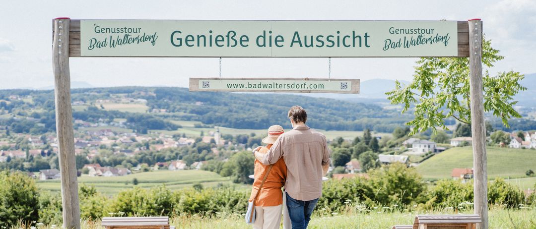 Zwei Menschen stehen auf einer Terrasse mit Blick auf die Landschaft und betrachten die Aussicht. Die Terrasse hat Holzbänke. Ein Schild mit der Aufschrift 'Genieße die Aussicht' hängt oben.