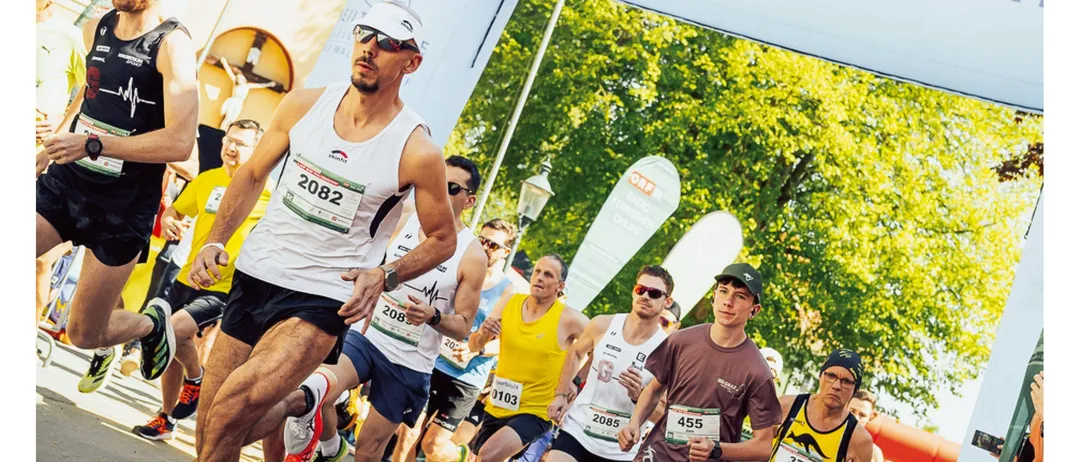 Läufer nehmen an einem Marathon teil und passieren ein Banner für Bad Waltersdorf. Das Banner trägt die Aufschrift 'www.badwaltersdorf.com'. Die Läufer tragen Nummern auf ihren Trikots. Der Slogan lautet 'Radio Steiermark Lauf. Jetzt anmelden.'