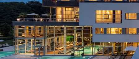 A modern hotel with a glass facade, illuminated at night. It features a large pool, a terrace with lounge chairs, and outdoor seating.