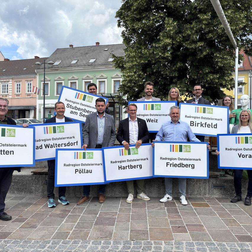 Eine Gruppe von Menschen steht draußen mit Schildern mit deutschem Text und posiert für ein Foto auf einem Stadtplatz. Gebäude mit Flaggen sind im Hintergrund.