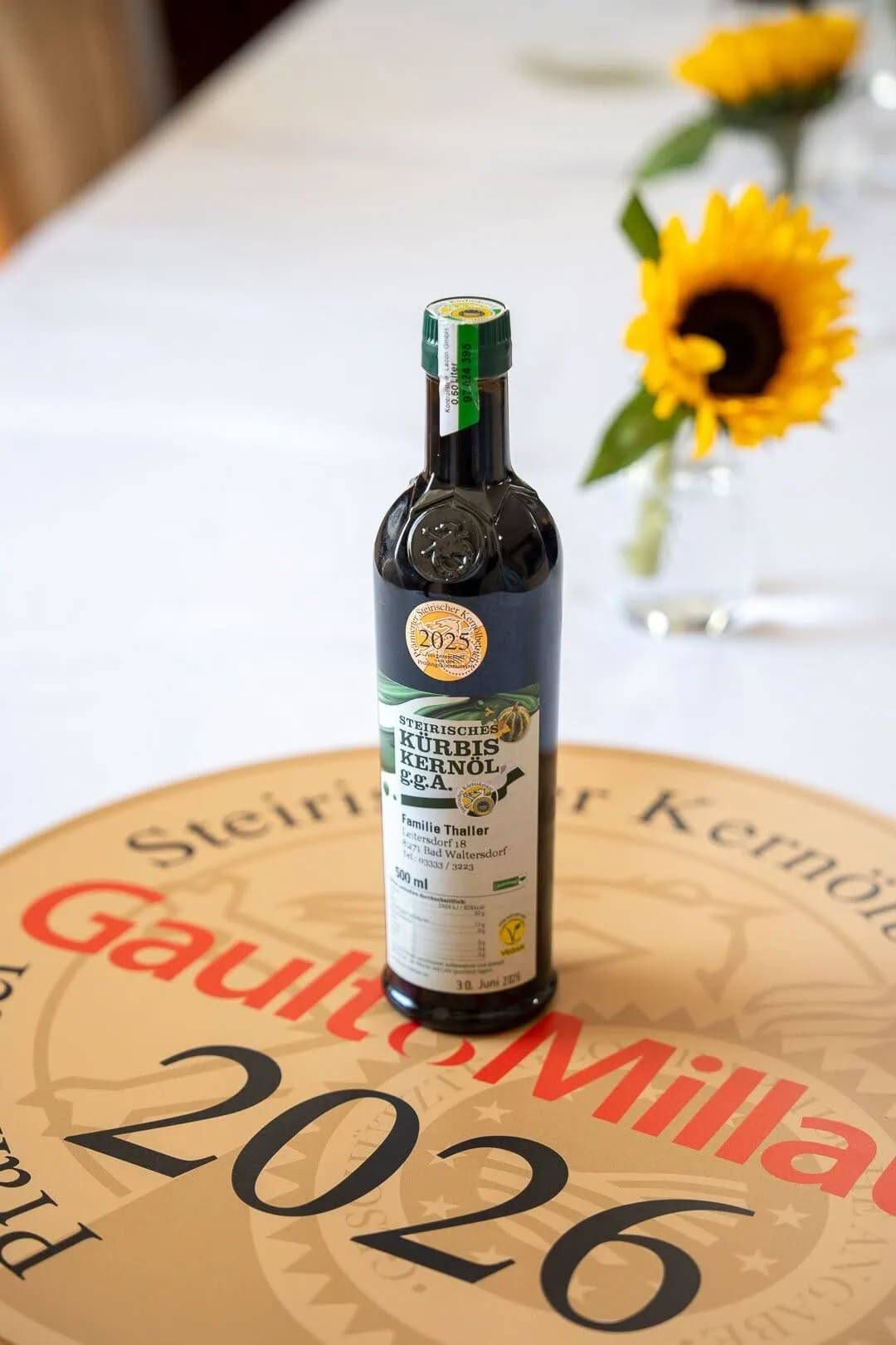 Eine Flasche Steirisches Kurbiskernöl auf einem kreisförmigen Etikett mit GaultMillau, mit einer Sonnenblume im Hintergrund.
