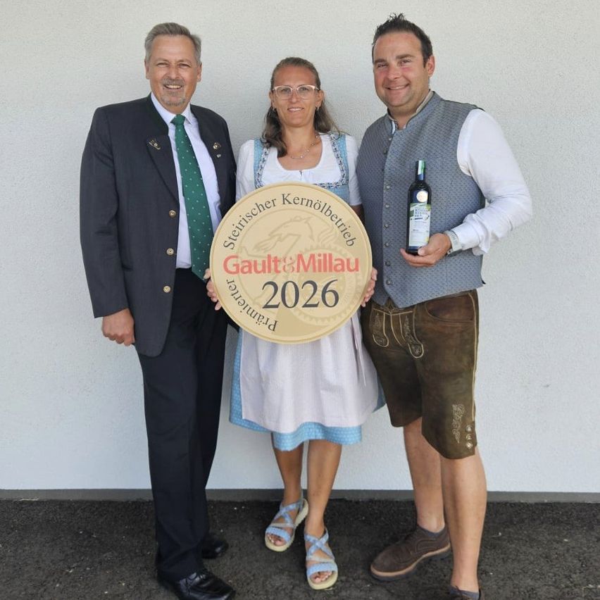 Drei Personen posieren lächelnd für ein Foto mit einer goldenen Plakette. Der Mann links trägt einen Anzug und eine Krawatte. Der Mann rechts hält eine Flasche Wein. Die Frau in der Mitte hält eine runde Plakette mit der Aufschrift Gault & Millau 2026.