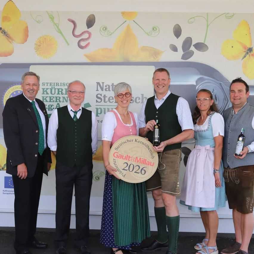 Fünf Personen stehen lächelnd in traditioneller Kleidung für ein Foto zusammen. Eine Frau hält ein kreisförmiges Schild mit der Aufschrift Gault&Millau 2026. Drei Männer und eine Frau halten Flaschen Wein in der Hand. Hinter ihnen befindet sich eine Wand mit einem Schmetterlings- und Blumendesign.