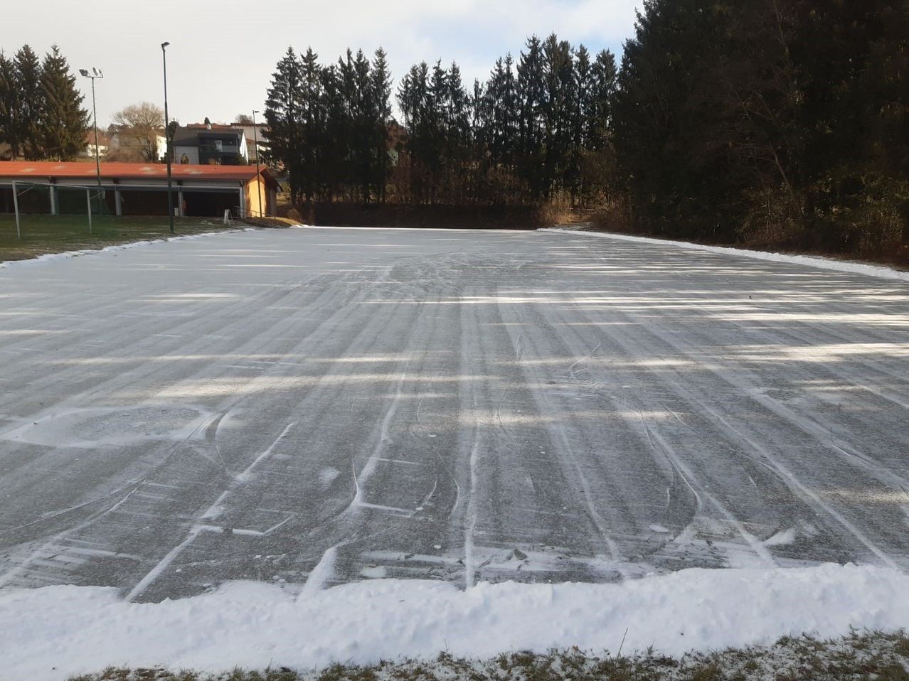 Eine breite, flache, vereiste Fläche erstreckt sich weit, gesäumt von Schnee und Bäumen in der Ferne. Im Hintergrund ist ein Gebäude mit einem roten Dach zu sehen.