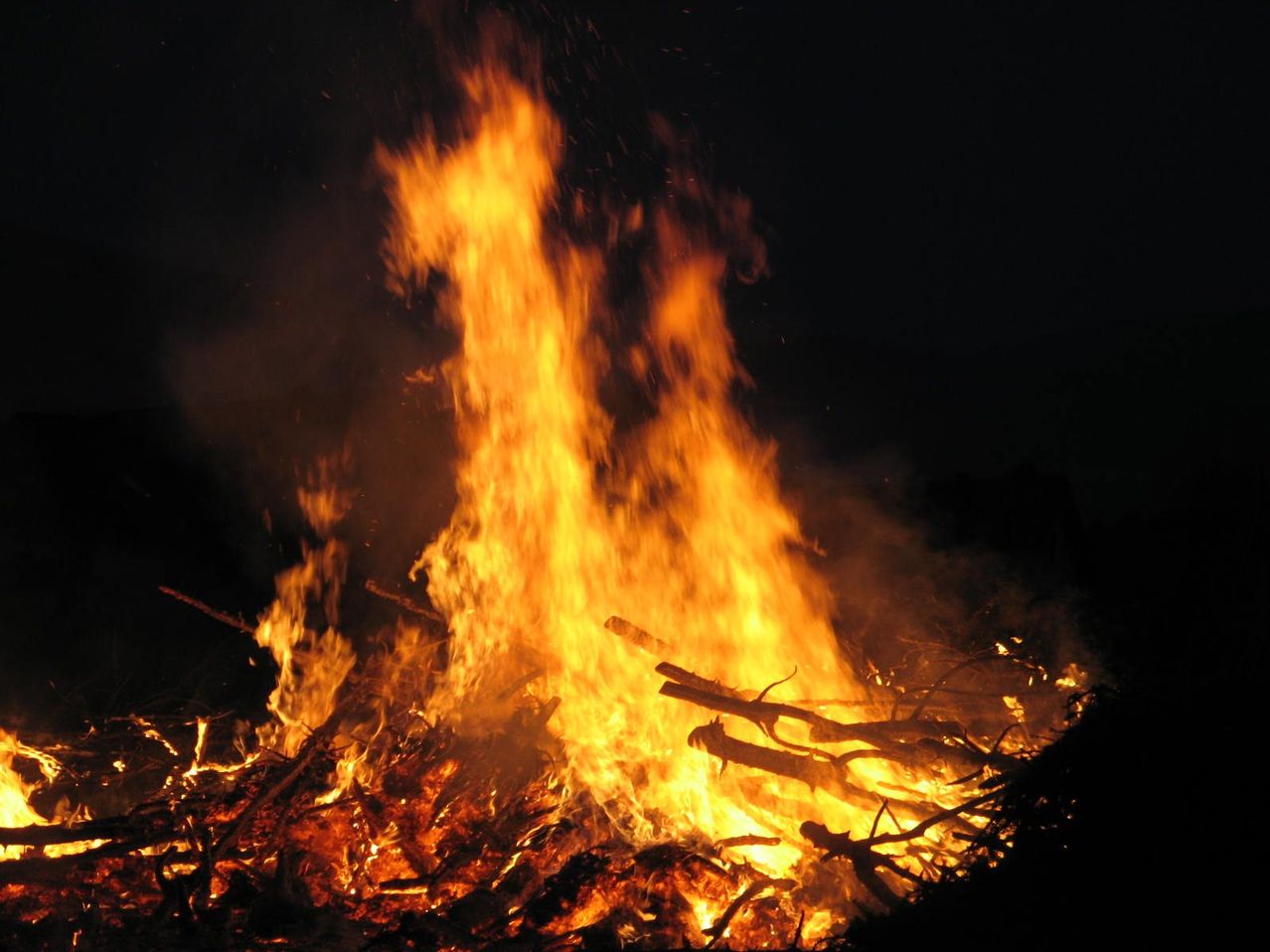 A bonfire burns brightly at night with flames reaching high and branches burning at its base.