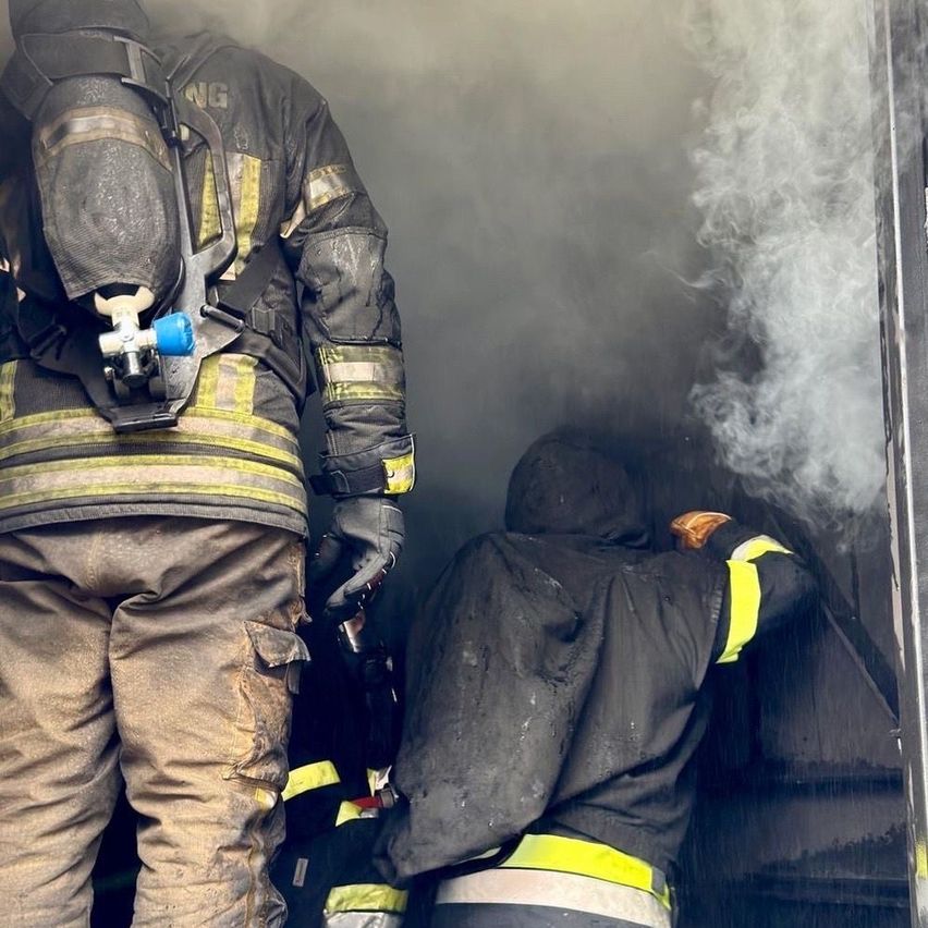 Feuerwehrleute in voller Ausrüstung verlassen ein rauchiges Gebäude. Ein Feuerwehrmann hat eine Maske auf, während ein anderer einen Feuerwehrbeil hat.
