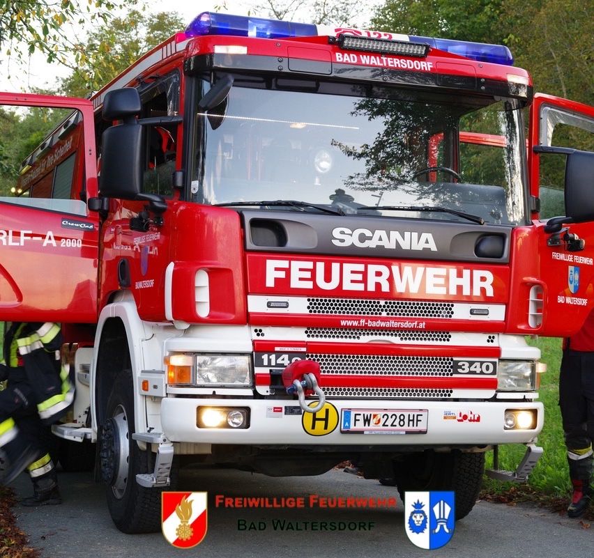 Ein roter Scania Feuerwehrwagen aus Bad Waltersdorf mit offenen Türen und einem Feuerwehrmann in der Nähe. Das Fahrzeug hat ein Nummernschild, Nummer 114, und ist auf der Straße geparkt.