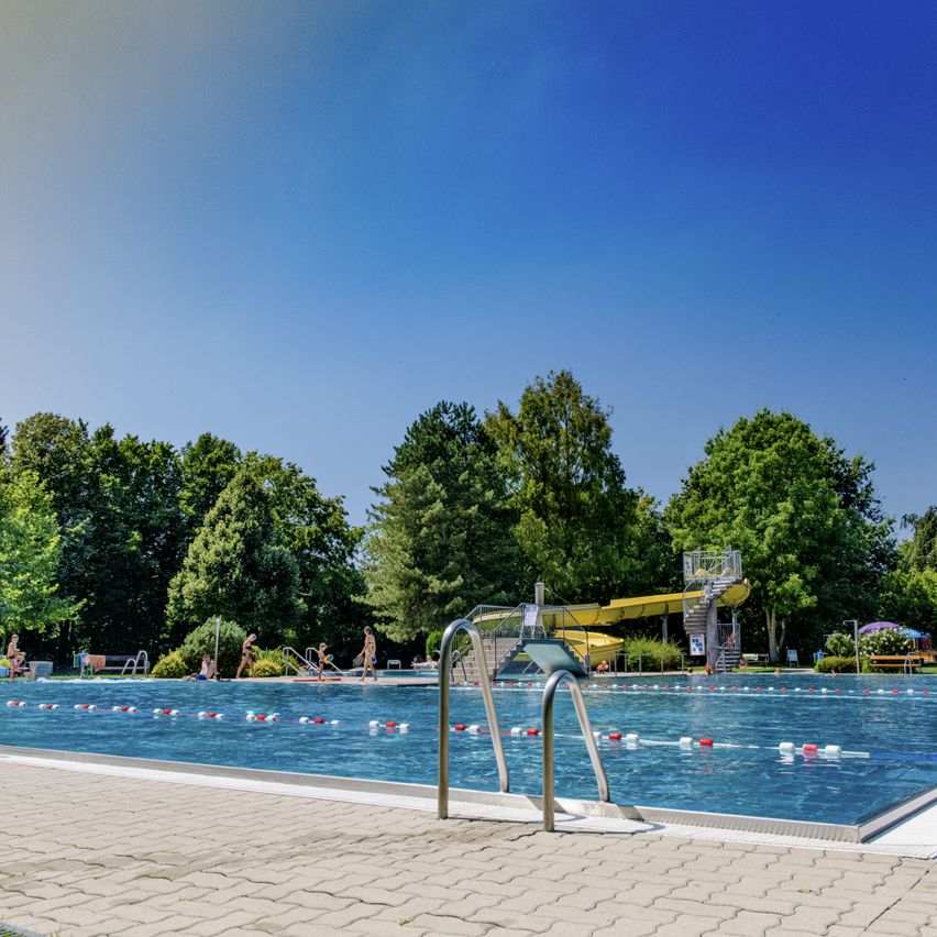 Ein Freibad mit blauem Wasser, Bahnmarkierungen, einem gelben Wasserrutsche und Menschen, die schwimmen und herumlaufen, vor dem Hintergrund üppiger grüner Bäume und eines klaren blauen Himmels.
