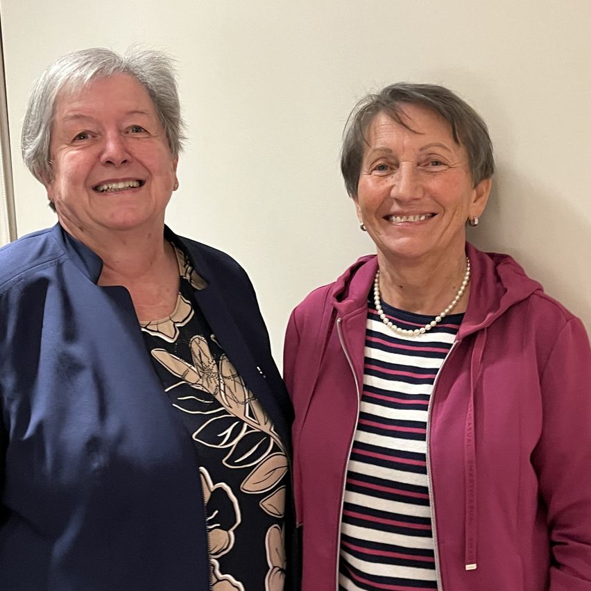 Two women stand close together, smiling. The woman on the left wears a blue blazer, and the woman on the right wears a pink jacket with a pearl necklace.