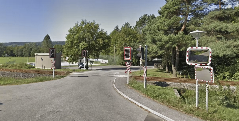 A road with a railway crossing signal, with two red traffic lights, a mirror, and a building on the left. Trees line both sides of the road.