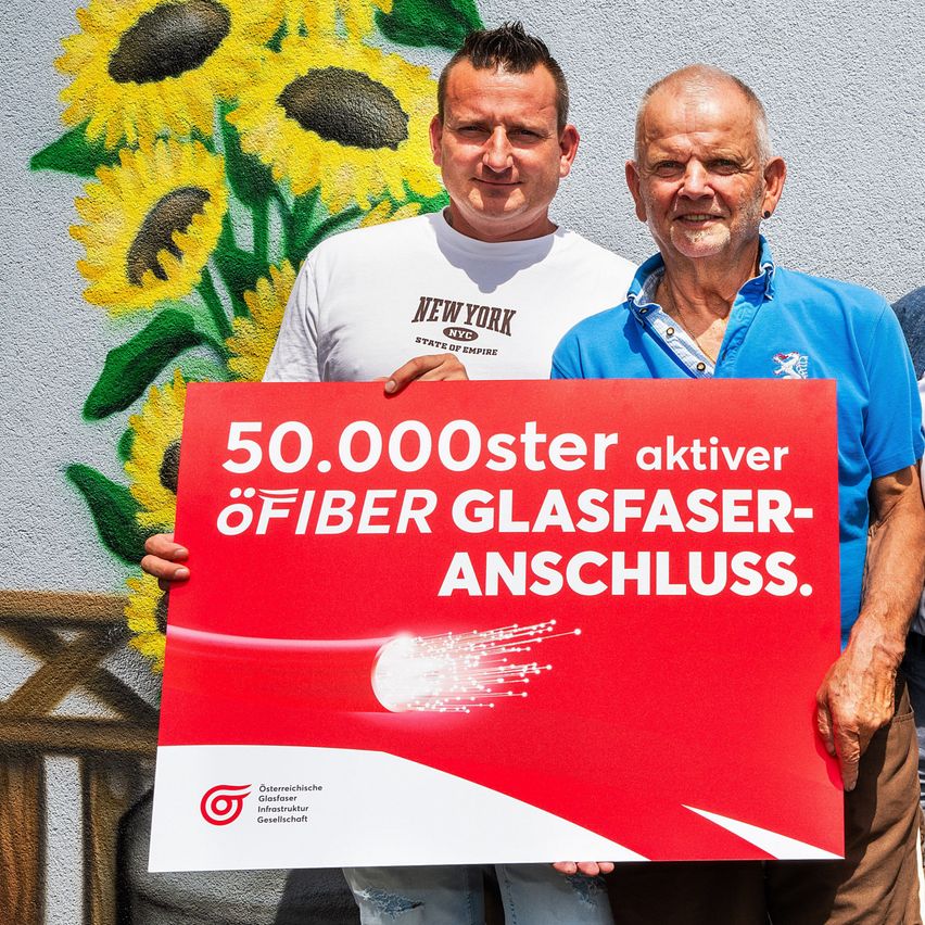 Zwei Männer stehen vor einer Sonnenblumenwand und halten ein rotes Schild mit der Aufschrift '50.000ster aktiver Glasfaser-Anschluss'. Der Mann auf der linken Seite trägt ein weißes 'New York'-T-Shirt, während der Mann auf der rechten Seite ein blaues Poloshirt trägt.