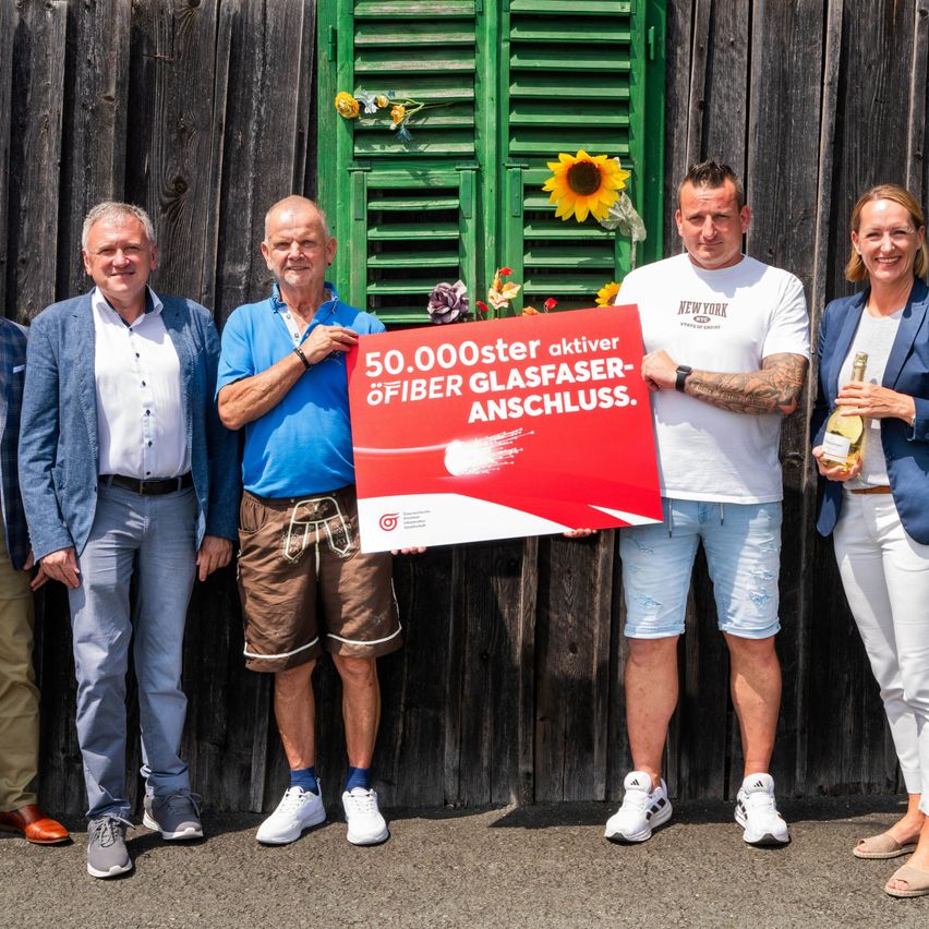 Fünf Männer und eine Frau stehen vor einer Holzwand mit einem grünen Fensterladen. Sie lächeln und halten ein rotes Banner mit dem Text '50.000 aktive Glasfaseranschlüsse'. Der Mann auf der rechten Seite hält eine Flasche.