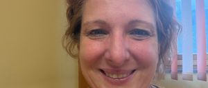 Eine Frau mit kurzen Haaren lächelt in die Kamera, trägt ein senffarbenes Oberteil und eine silberne Halskette. Sie befindet sich in einer Innenumgebung mit einer gelben Wand und einem Fenster mit Jalousien hinter ihr.