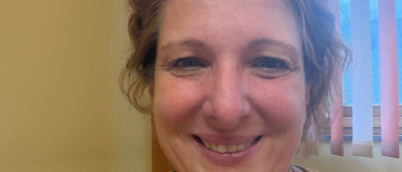 Eine Frau mit kurzen Haaren lächelt in die Kamera, trägt ein senffarbenes Oberteil und eine silberne Halskette. Sie befindet sich in einer Innenumgebung mit einer gelben Wand und einem Fenster mit Jalousien hinter ihr.