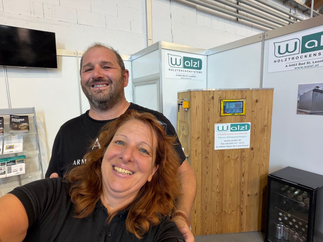 Ein Mann und eine Frau lächeln und posieren für ein Foto in einem Raum mit einem hölzernen Stand, der mit 'Holztrockensystem' beschriftet ist.