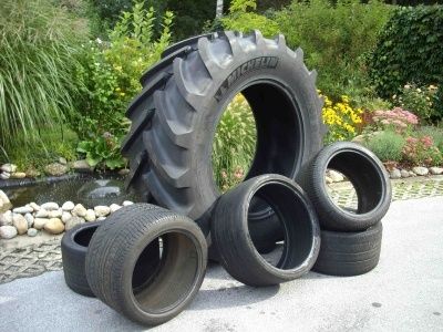 Eine Sammlung schwarzer Traktor- und Autoreifen wird auf einer Betonfläche in einem Gartenbereich ausgestellt. Dahinter befinden sich Pflanzen, Blumen und ein Teich.