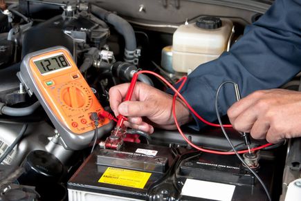 Ein Mechaniker arbeitet an einer Autobatterie, hält ein Multimeter und verbindet rote und schwarze Kabel. Das Multimeter zeigt 27,8 Volt an.