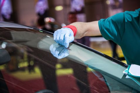 Eine Person in Handschuhen arbeitet an der Windschutzscheibe eines Autos, möglicherweise eine Rissreparatur. Die Windschutzscheibe ist leicht gebogen, und die Reflexion der Person ist sichtbar.