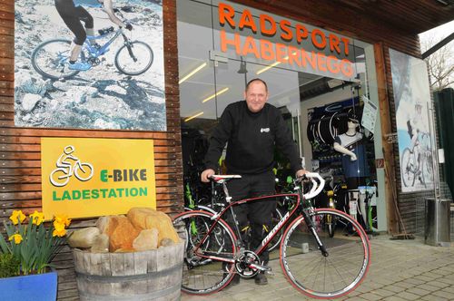 Ein Mann in einem schwarzen Outfit steht vor einem Geschäft namens Radsport Haberneg, mit einem Fahrrad in den Händen. Das Geschäft zeigt ein Schild für E-Bikes.