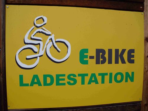Ein gelbes Schild zeigt eine weiße Illustration einer Person, die ein Fahrrad fährt. Darunter steht in grünen und schwarzen Buchstaben 'E-BIKE LADESTATION'.