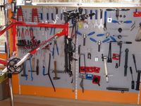Ein rotes Fahrrad hängt an einer Wand mit verschiedenen Werkzeugen. Die Werkzeuge umfassen Schraubenschlüssel, Schraubendreher und Hämmer. Das Fahrrad ist mit 'Simplon' beschriftet.
