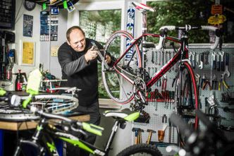 Ein Mann repariert ein Fahrradrad in einer Garage mit Werkzeug und Ausrüstung an der Wand. Er hält ein Werkzeug in der Hand und das Fahrradrad hängt an einem Ständer.