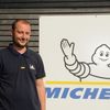 Ein Mann in einem dunkelblauen Hemd steht neben einem weißen Schild mit einem Michelin-Logo und einer gezeichneten Figur, die winkt.