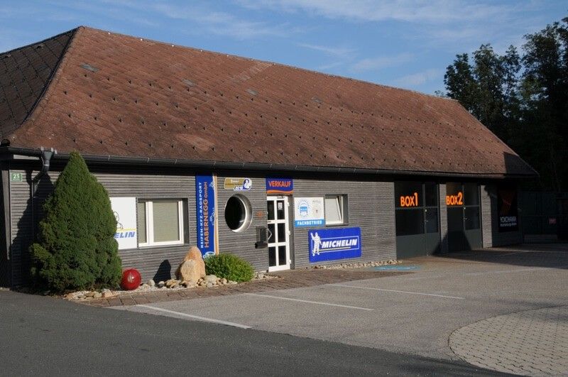Das Gebäude hat ein braunes Dach und einen Parkplatz davor. Der Eingang hat eine Glastür mit einem runden Fenster. Das Schild sagt Michelin.