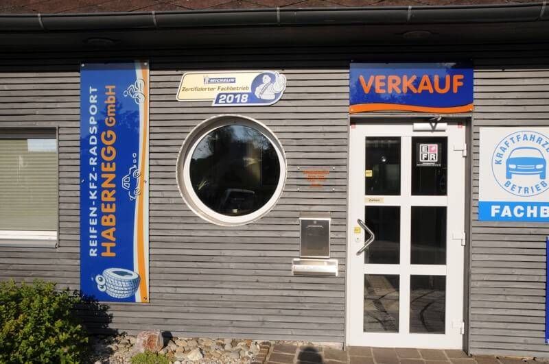 Das Gebäude hat ein Schild mit der Aufschrift Verkauf und ein Fenster, das ein Fahrzeug reflektiert. Es gibt eine Tür mit einem Griff und ein kleines Schild daneben. Das Gebäude hat auch ein blaues Banner mit Reifen-Kfz-Radsport und einem Logo.