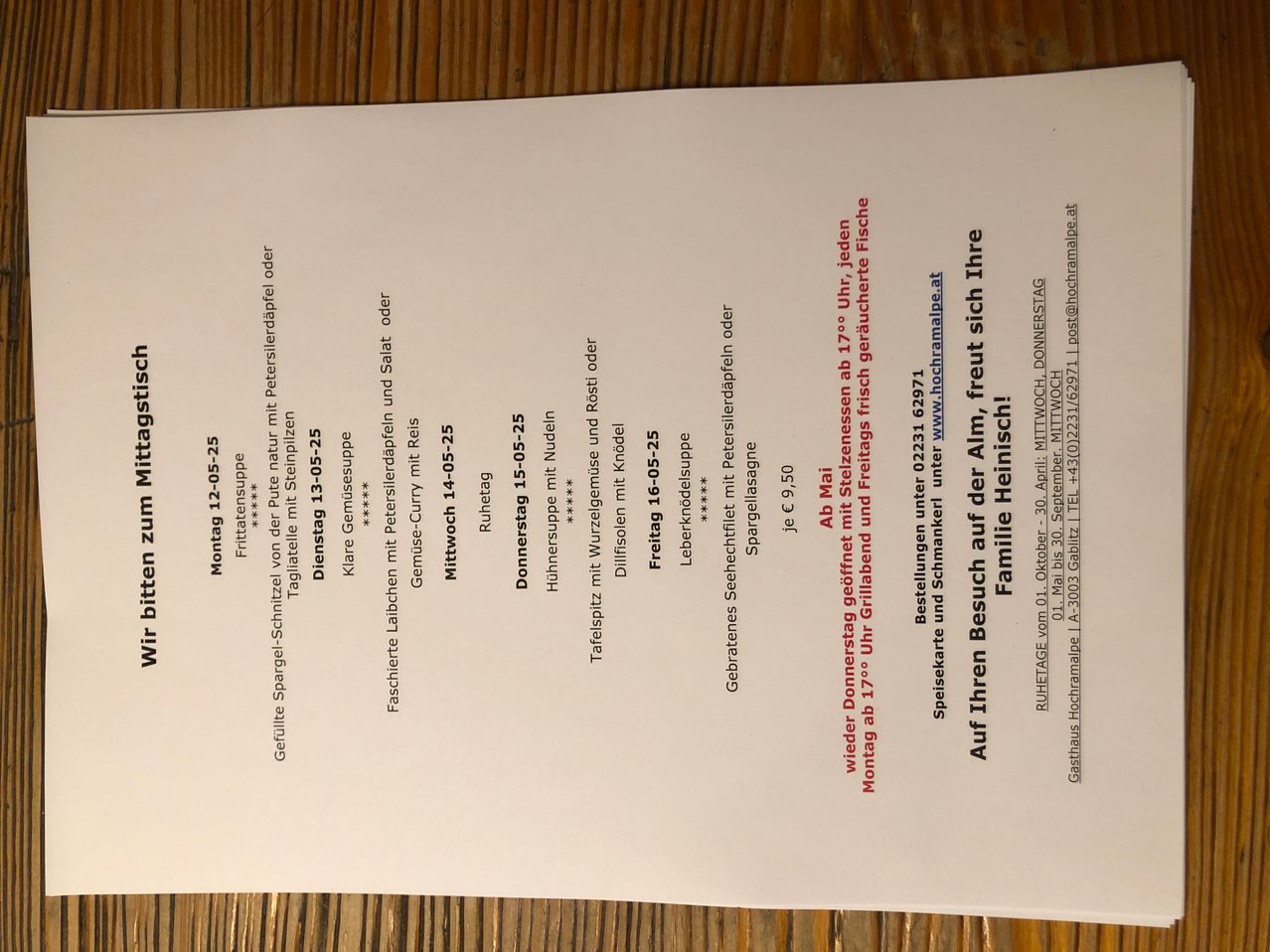 Ein weißes Blatt Papier zeigt eine Speisekarte oder einen Zeitplan mit verschiedenen Daten und Artikeln, alles in Rot und Schwarz geschrieben. Die Artikel umfassen Gerichte und Preise, wobei die Daten von Montag 12-05-25 bis Freitag 16-05-25 reichen. Die Preise sind in Euro angegeben. Der Text ist leicht verschwommen.