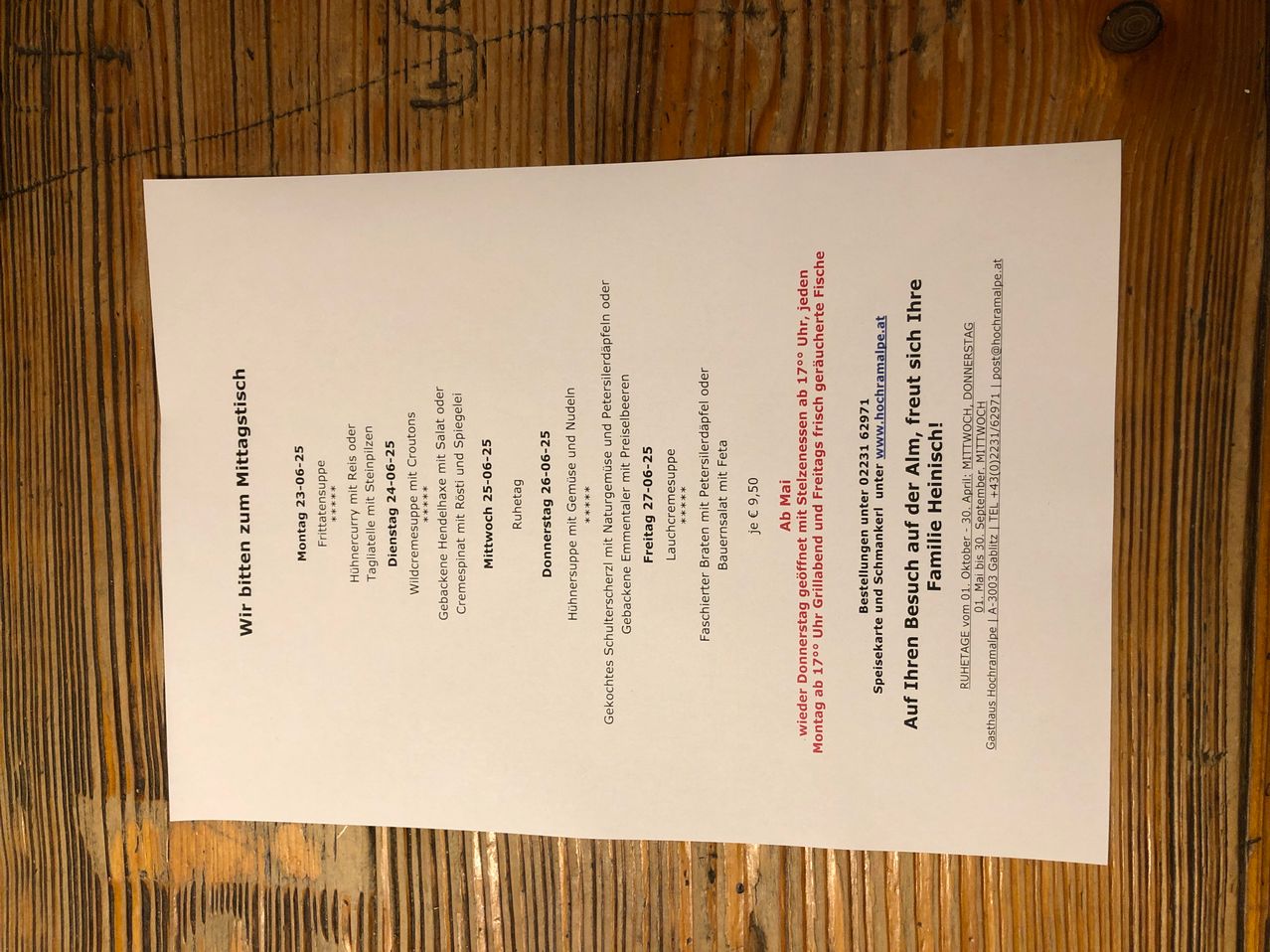 Ein weißes Dokument auf einer hölzernen Oberfläche mit Text in roten und schwarzen Schriftarten. Es scheint sich um eine Speisekarte oder einen Zeitplan zu handeln, mit Daten, Namen und Preisen. Das Dokument enthält Namen wie 'Donnerstag 26-25', 'Freitag 27-25' und 'Montag 23-06'. Die Preise sind im Format '12,90 Euro' aufgeführt. Der Text ist in einer Rasterstruktur organisiert.