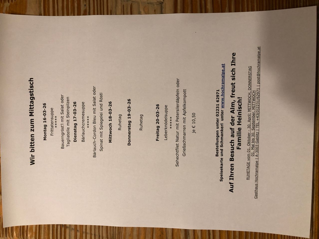 Ein weißes Dokument auf einer Holzoberfläche listet wöchentliche Mahlzeitenoptionen, Zutaten und Preise auf. Es enthält Mahlzeiten von Montag bis Freitag, wobei jeder Tag ein anderes Hauptgericht, Suppe und Dessert sowie einen Preis von 10 Euro hat.