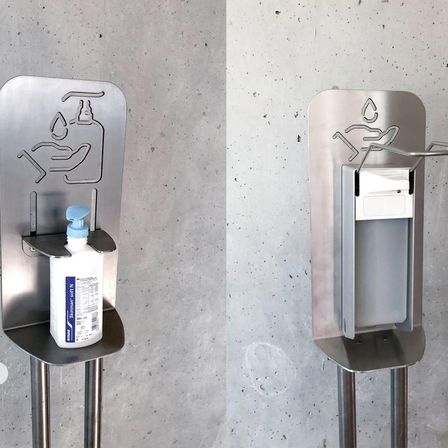 Ein Edelstahl-Händedesinfektionsmittelhalter mit einer Flasche und einem Spender an einer Betonwand.