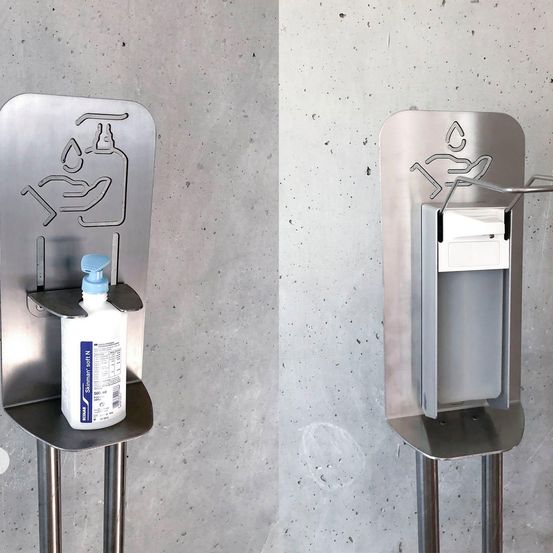 Ein Edelstahl-Händedesinfektionsmittelhalter mit einer Flasche und einem Spender an einer Betonwand.