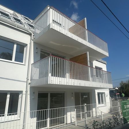 Ein modernes weißes Apartmentgebäude mit mehreren Balkonen und Glastüren, vor einem klaren blauen Himmel.