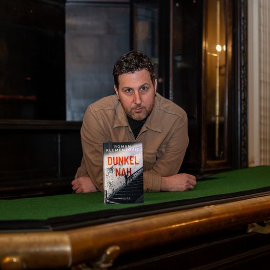 Ein Mann lehnt sich über einen Tisch mit einem Buch namens 'Dunkel Nah' vor sich. Er trägt ein braunes Hemd und einen Schnurrbart. Der Tisch hat eine grüne Oberfläche und goldene Einfassung. Hinter ihm befindet sich ein dunkler Raum mit einer Glastür.